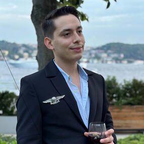 Cenk Topuz in a suit looking ahead with a smile while holding glass in a nature background